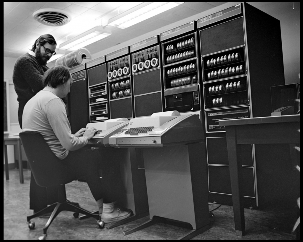 Undated photograph, probably circa late nineteen sixties of a Unix developers Ken Thompson and Dennis Ritchie at a DEC PDP 11 computer running Unix. In the left of the photo, Ritchie is standing and watching Thompson, who is sitting at one of two teletype terminals used as consoles to interface with the computer.