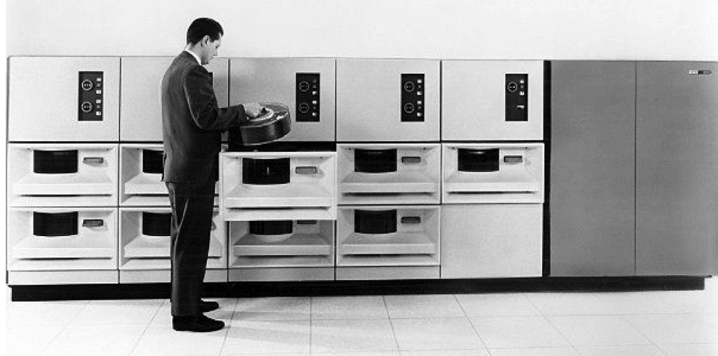 A black and white photo showing a row of IBM 2314 disk drives with a man mounting a removable disk pack.
